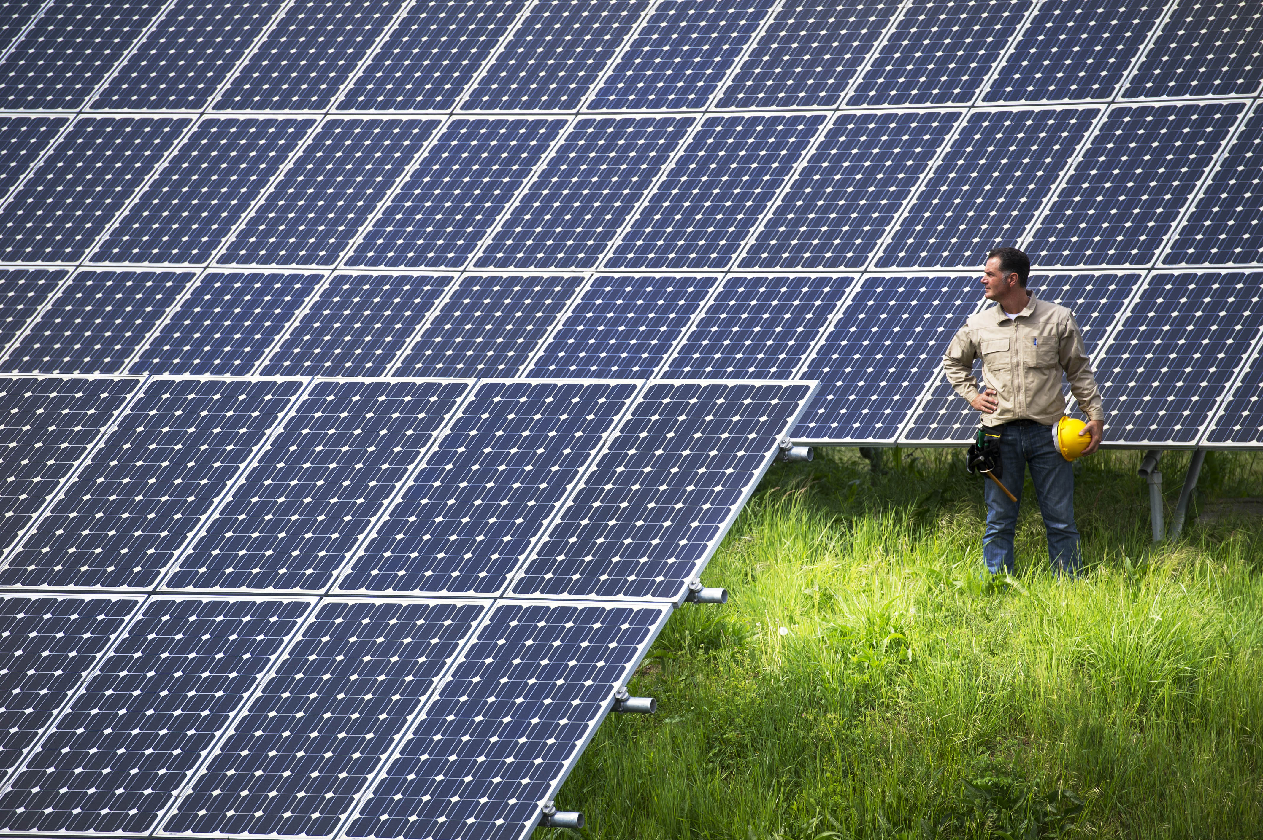 Photo of a man holding a hardhat standing to the right of a large array of newly installed solar panels.